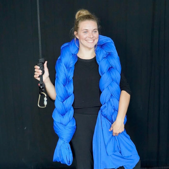 A young white woman stands smiling with a large braid of blue fabric draped over her shoulders. In one hand she holds a sturdy black rope with a carabiner tied to one end.