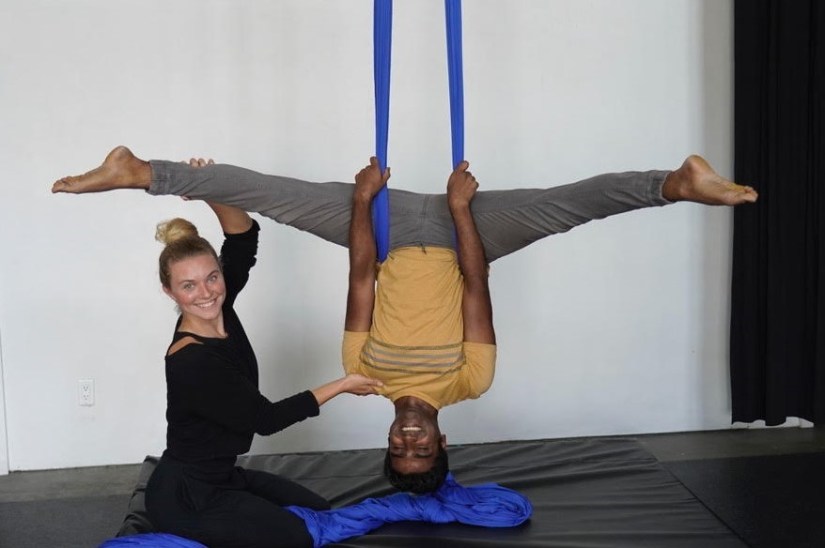 A young white woman supports a young brown man as he holds an upside down straddle pose on blue aerial fabric. Both people are beaming widely.