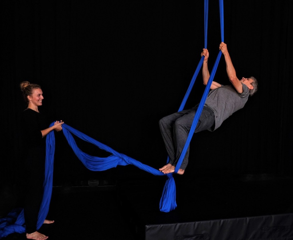 A young white woman holds the ends of blue aerial fabric as an older white person swings on the fabric.
