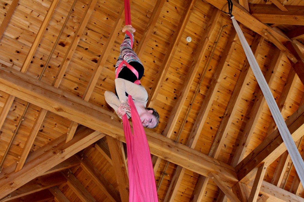 A young white teenager is suspended upside down on two pieces of pink aerial fabric. She is beaming. 