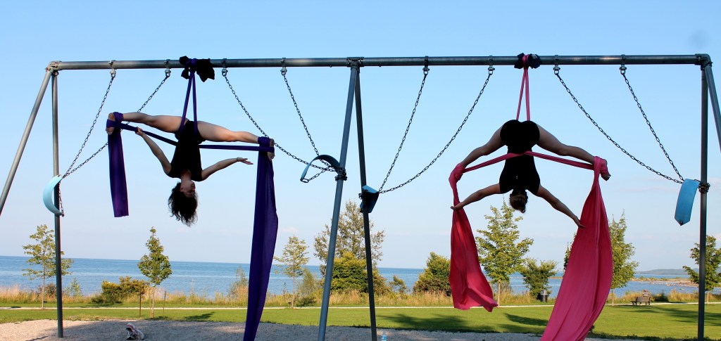 Two white teenage girls hang upside down on fabric draped over a swingset. 