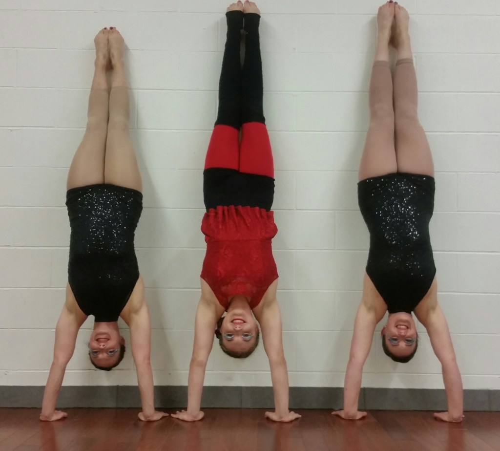 Three white women are smiling doing handstands against a wall. Two of the women are wearing black sparkly biketards with nude tights and bare feet. The midle woman is wearing a costume with a bright red top and leggings,overlayed with black shorts and legwarmers that cover her knees.  