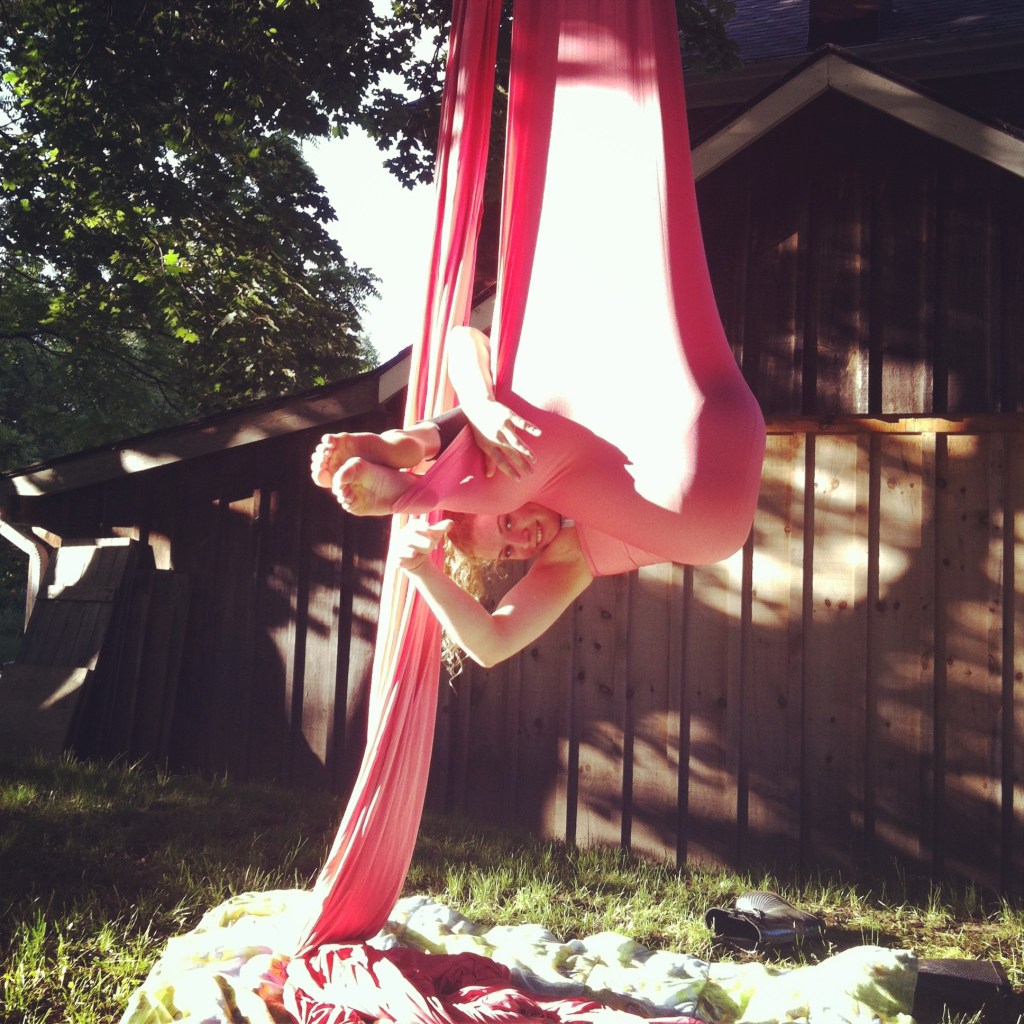A young white woman peeks out from within a hammock of pink aerial fabric. She looks happy. Behind her is a shed, the photo appears to be taken in a backyard. Underneath her there is nothing but a blanket. 