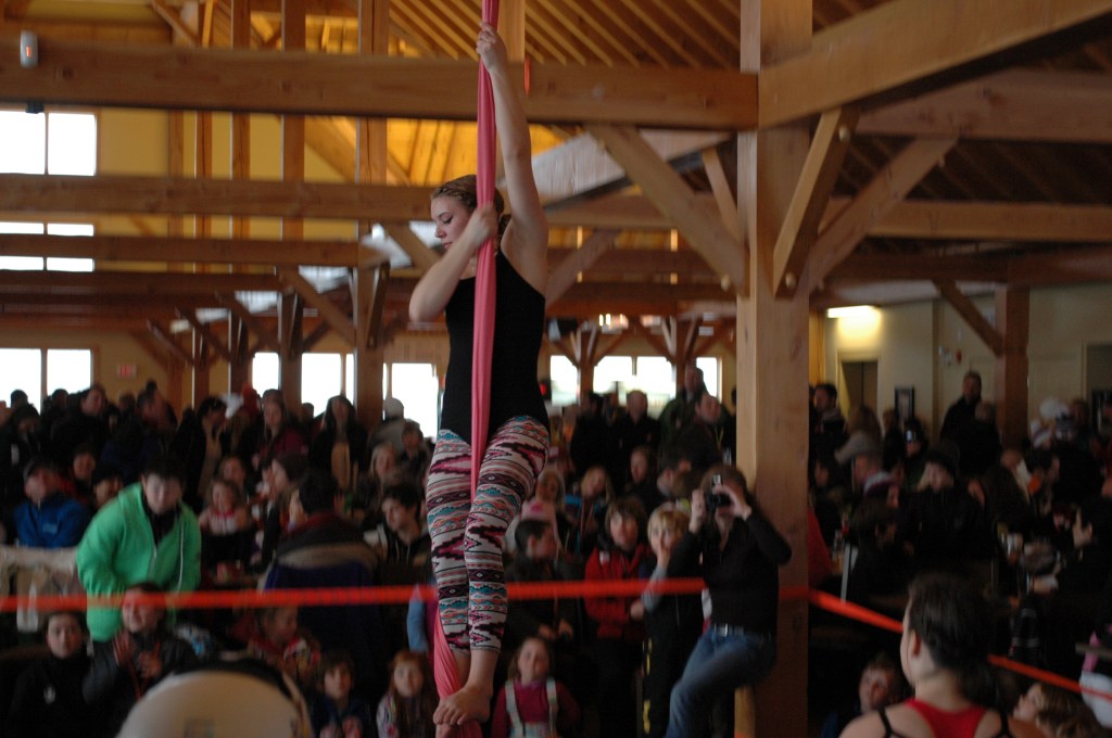 A young white woman slides down long pink aerial fabric. Behind her there is a photographer and a large crowd of people. 