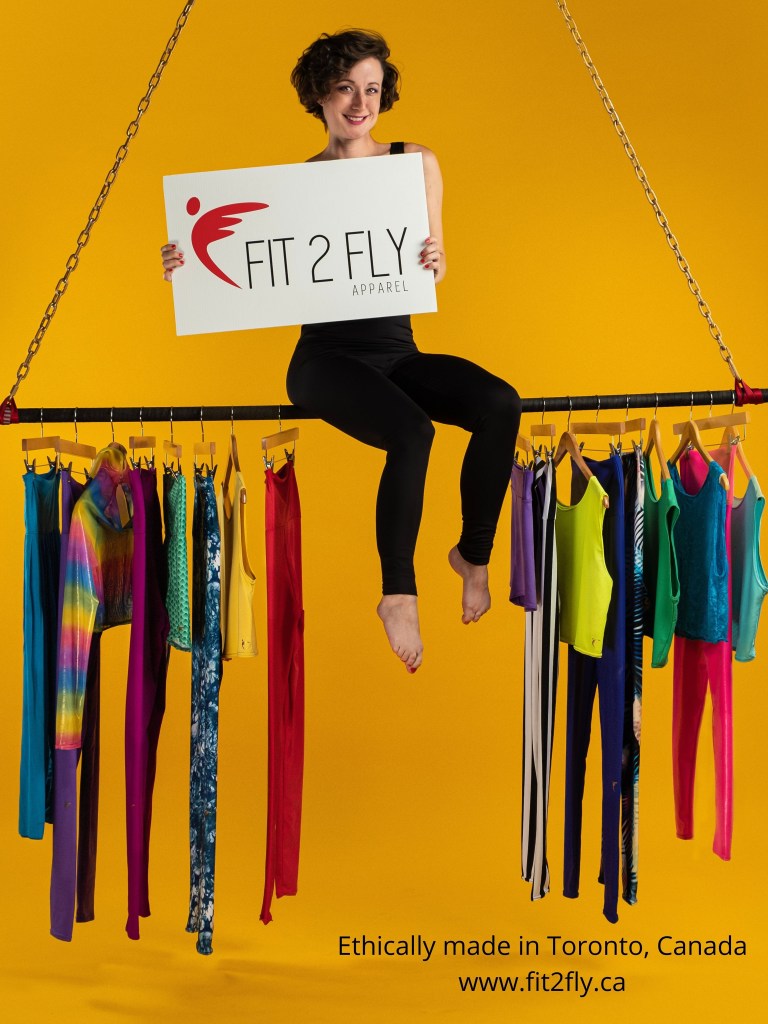 A young white woman sits on top of a suspended metal bar against a yellow background. She is holding a sign that reads Fit 2 Fly Apparel". Below her, an assortment of colourful leggings and tops are hanging off the bottom of the bar. 