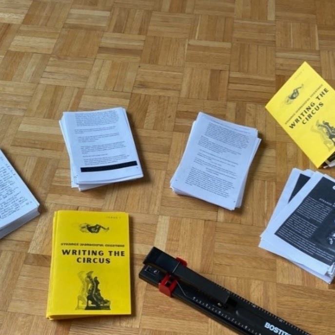 Pieces of folded paper with text on them are arranged orderly against a wooden floor. One of the piles of paper stands out, a yellow page with blurry images of humans and bold text that reads "Writing the Circus".