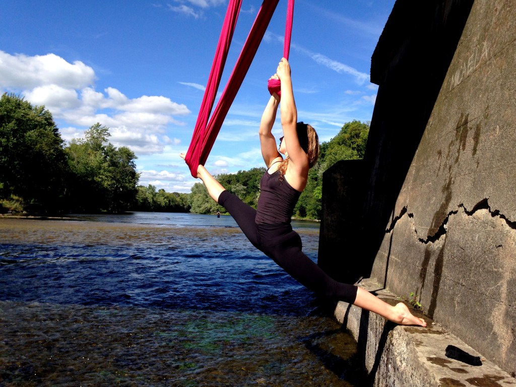 A young white woman hangs on pink aerial fabric over a calm stream. She is looking into the distance. 