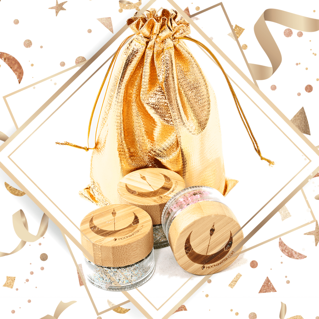 A shiny gold bag is featured against a white and gold background. Below the bag sit three small containers, clear on the bottom with a wooden top.  Each small jar full of a different colour of glitter, white, green, and pink. The lids of the jars have a crescent moon on them.
