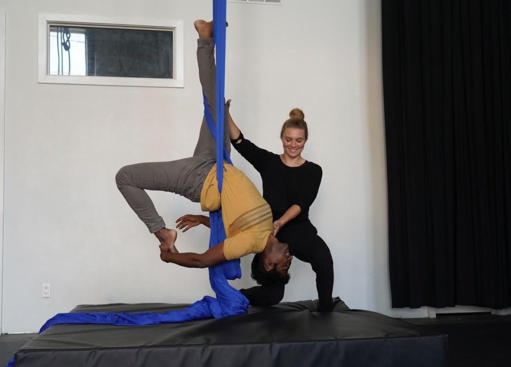 A young white woman, supports a young brown man as he hangs upside down on blue aerial fabric. Both people are smiling.  