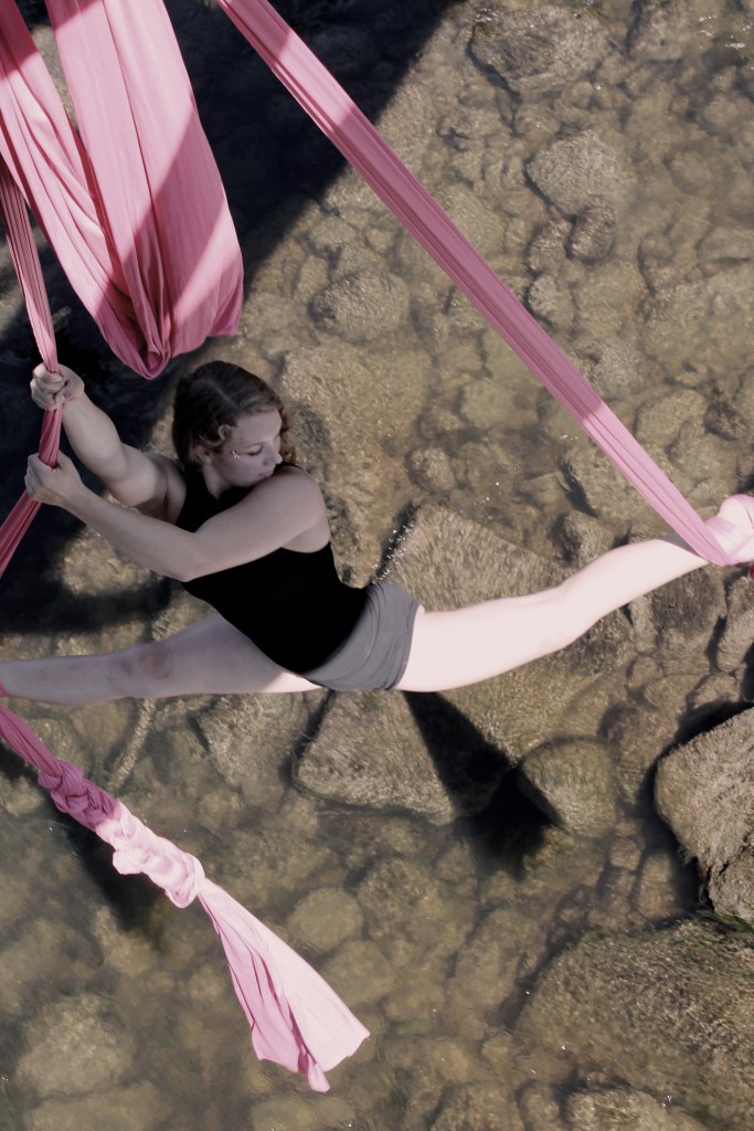 A young white woman is suspended over a calm river hanging off of two pink pieces of aerial fabric. 