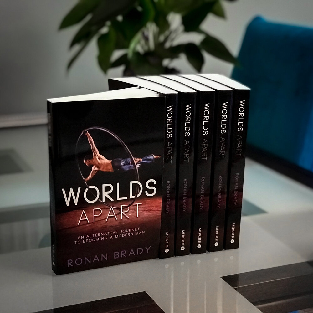 A stack of books stands upright on a shiny table. The front cover is black and sleek with white text reading "Worlds Apart: an alternative journey to becoming a modern man. By Ronan Brady." Behind the text a muscular white man in only blue jeans spins on a large metal ring known as a Cyr wheel.  